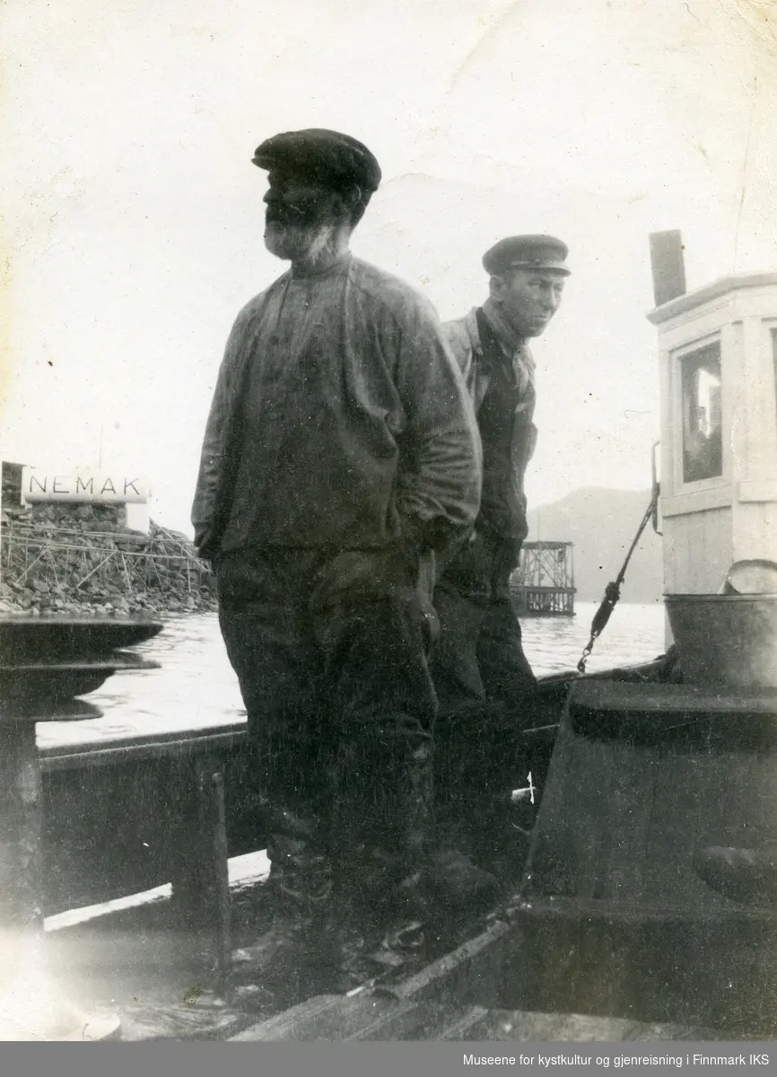 To menn i en båt på Honningsvåg havn. I bakgrunnen fiskehjeller, en NEMAK-tank og til høyre en nothjell. 1930-årene.