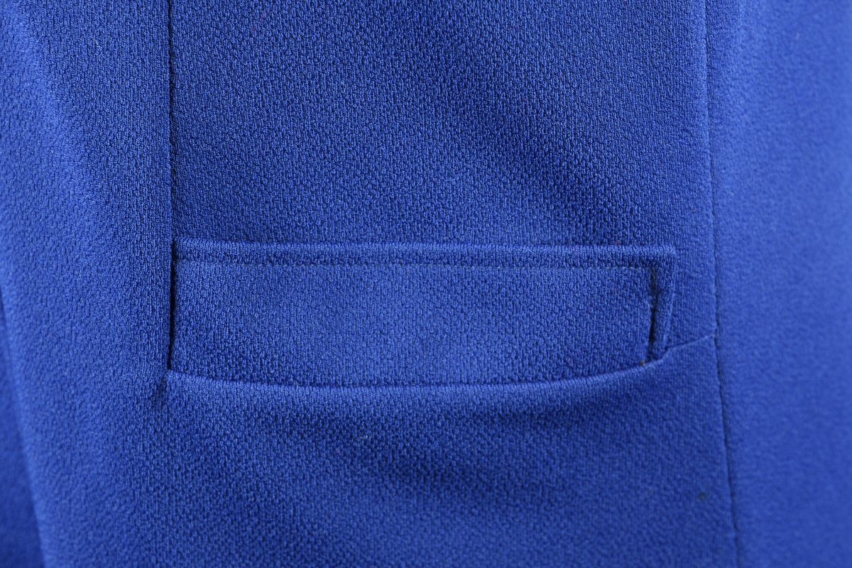 Selekjole av blå trikot. Kjolen har spensel i ryggen som er festet med to knapper som er trukket i kjolens stoff. Kjolen lukkes i front med syv knapper som også er trukket i kjolens stoff.