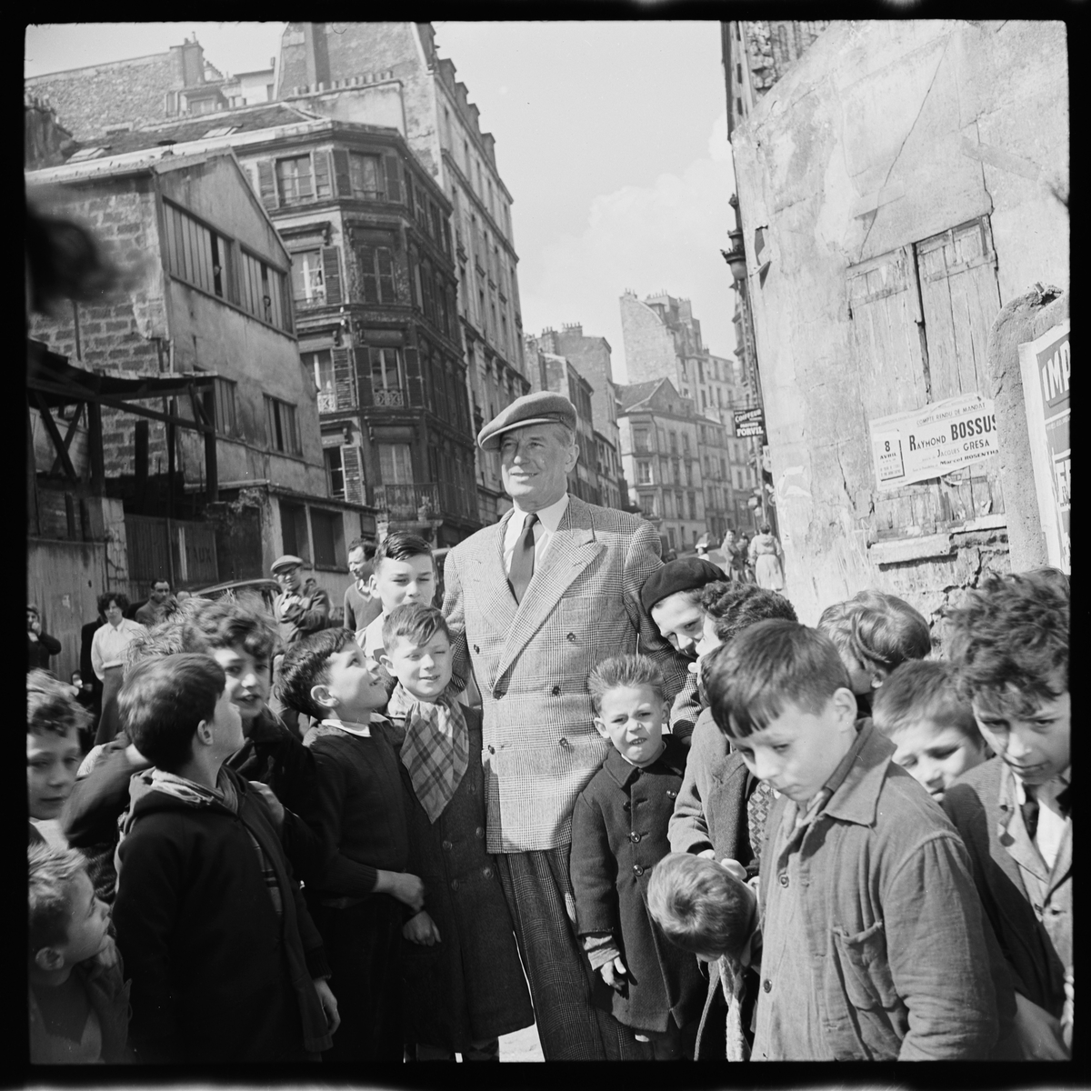 Maurice Chevalier og en flokk med unger. I bakgrunnen bygninger. 