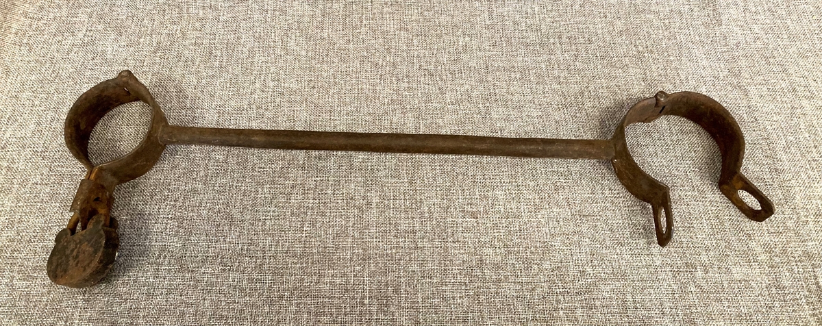 To leddede håndjern forbundet med en lang jernbolt. Begge jernene kan låses med hengelås. Den ene hengelåsen mangler.
