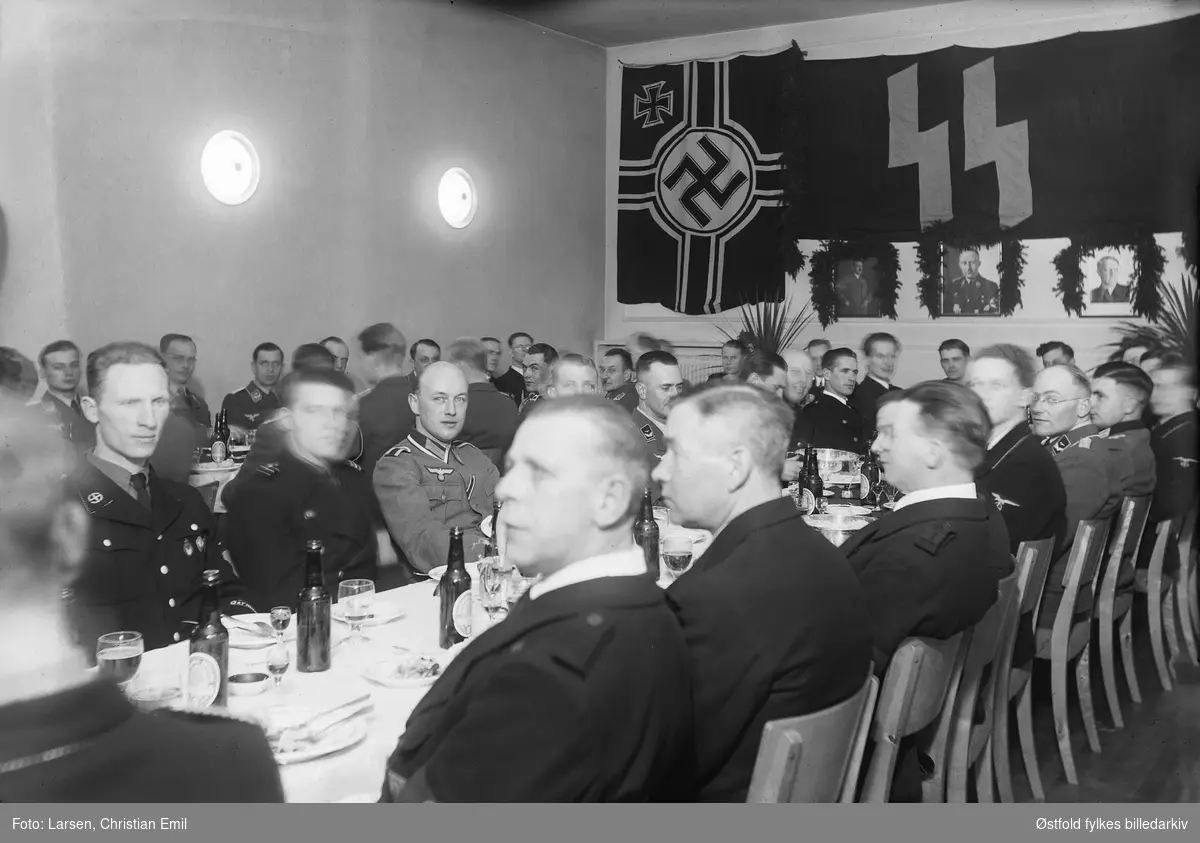 Medlemmer av Germanske SS Norge og tyske soldater samlet til fest. Øl og dram på bordet (pils fra Sarpsborg bryggeri). I bakgrunn naziflagg, og SS fane og portretter av Hitler, NN, Vidkun Quisling som er pynta med granbar.
Kan være fra Festiviteten i Sarpsborg (Olavs Hall).