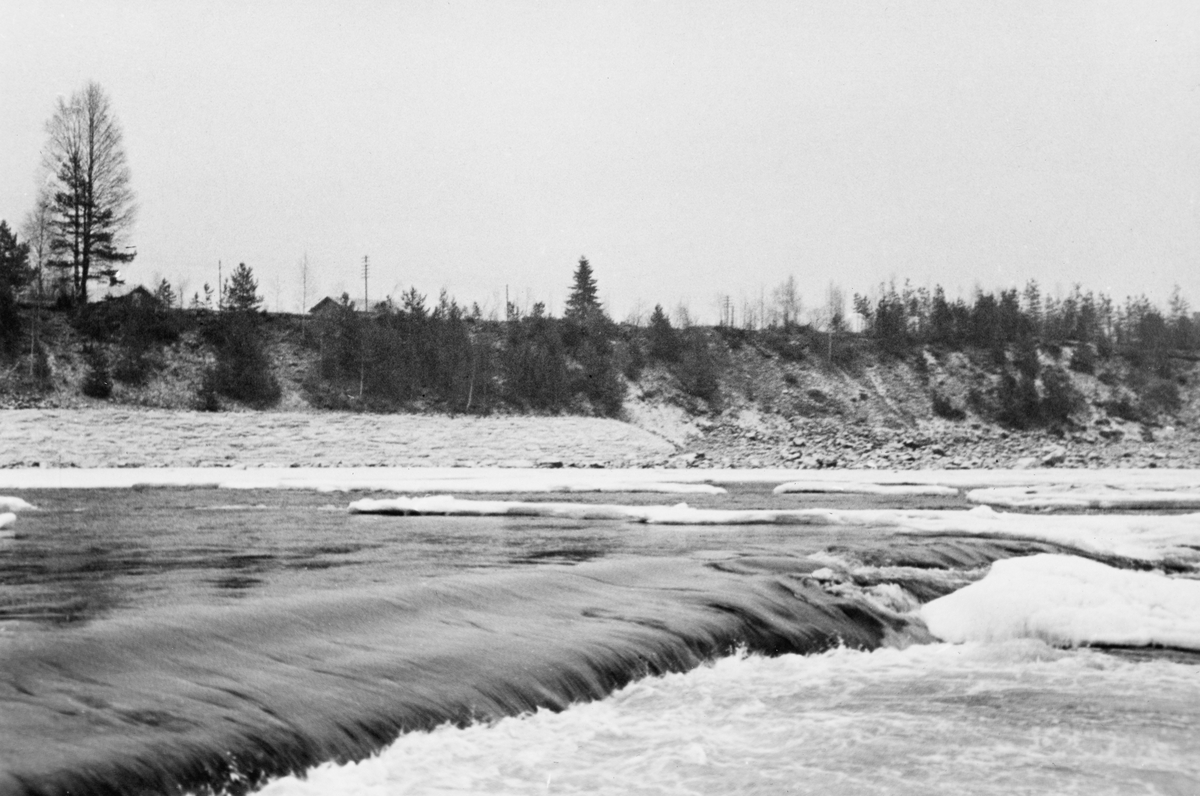 Fra Glommas løp ved Skjefstadfossen i Heradsbygda i Elverum i Hedmark. Fotografiet ble tatt seinhøstes i 1933, etter at de stilleflytende delene av elveløpet var dekt av is med et tynt snølag, men med ei råk ved en liten terskel i elveløpet i forgrunnen. I bakgrunnen ser vi en bratt, steinete elveskråning og på elvesletta bakenfor skimter vi røstene på et par bygninger på et lite gardsbruk.