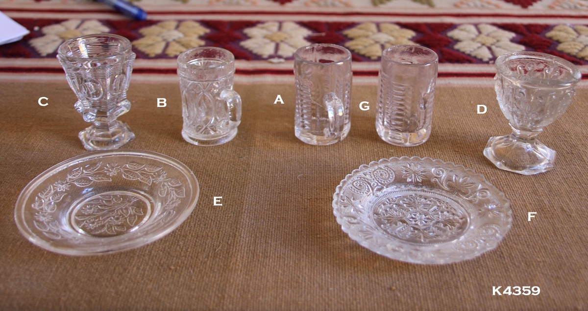 Glassanretning:
Innhold: 4 drammeglass, 1 eggeglass og 2 smørskåler.
2 like drammeglass (a). Det ene mangler hank (g).
1 drammeglass på stett (c). Bunn 6-kantet regulær mangekant.
1 eggeklass (d). Bunn 8-kantet regulær mangekant.
1 smørskål med bladmønster (e).
1 smørskål med blondekant og mønster (f).