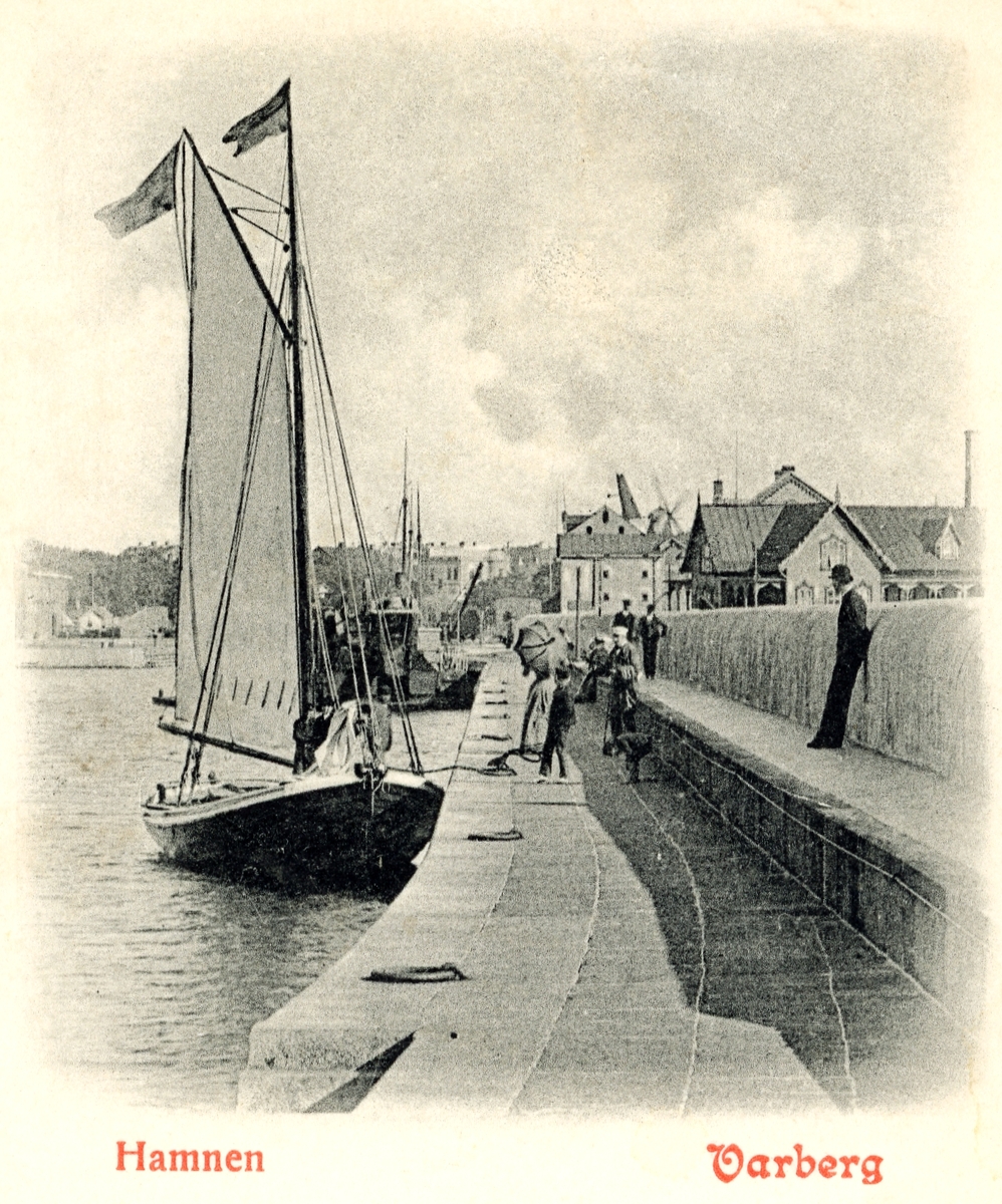 Brevkort (före 1904), "Hamnen Varberg". Vy från hamnpiren med en segelbåt i förgrunden, varmbadhuset till höger och bakom det Lundqvists spannmålsmagasin, sammanbyggt med hamnmagasinet varifrån rian skjuter upp.