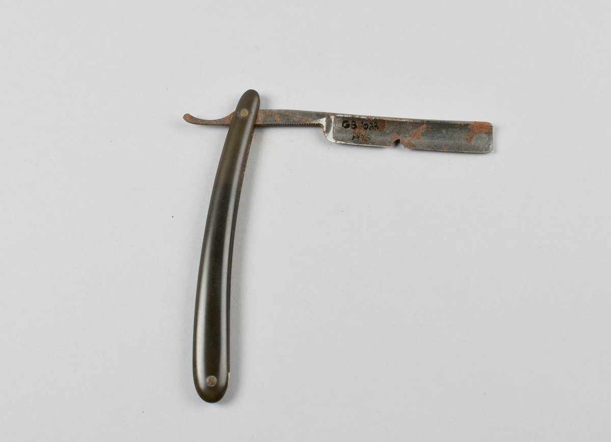 Sammenleggbar barberkniv med stålblad, skaft i svart celluloid. Et stykke hvit celluloid er innfelt bak på skaftet mellom de to svarte skaftdelene.
