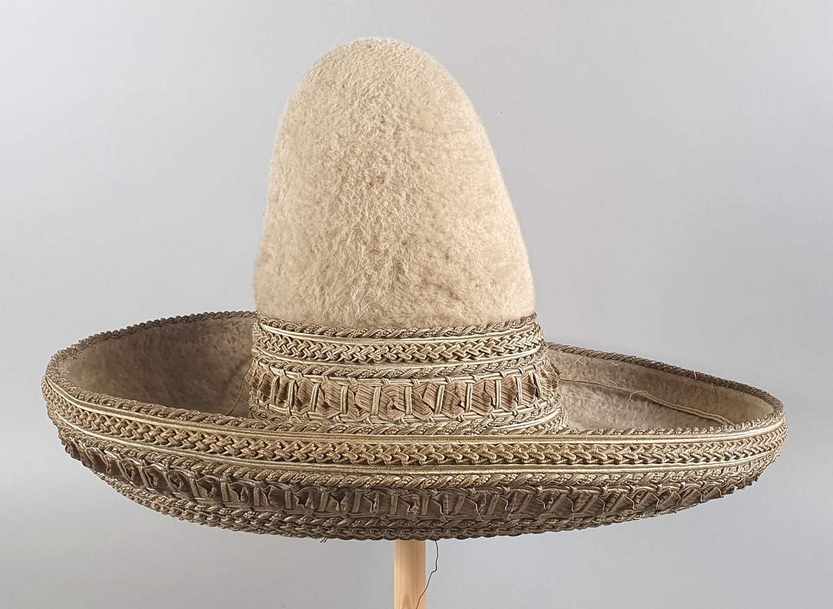 Beige (sauehvit) meksikansk hatt av børstet ull. Hatten har høy smal pull og bred brem som vender oppover. Flettede brede bånd av strå med sølvtråd rundt pullen på yttersiden og bremmen på undersiden. Smalt kantbånd ytterst på bremmen har nesten revnet helt av.