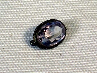 Oval genomskinlig slipad sten av något slag i ram av tunt flätat band av mässing. På baksidan en säkerhetsnål med vilken man fäster broschen på plagget.