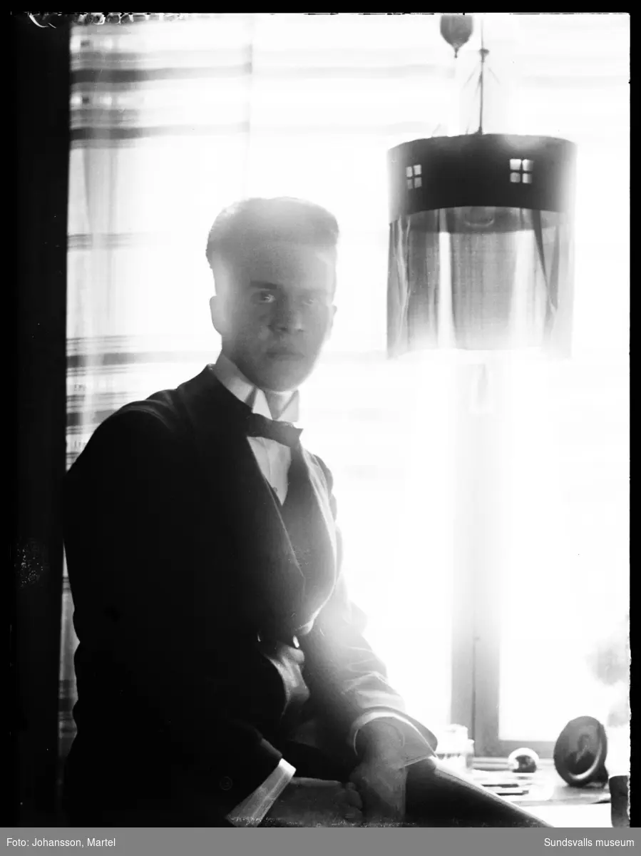 Porträttbild inomhus av en man som sitter i motljus intill ett fönster och en taklampa.