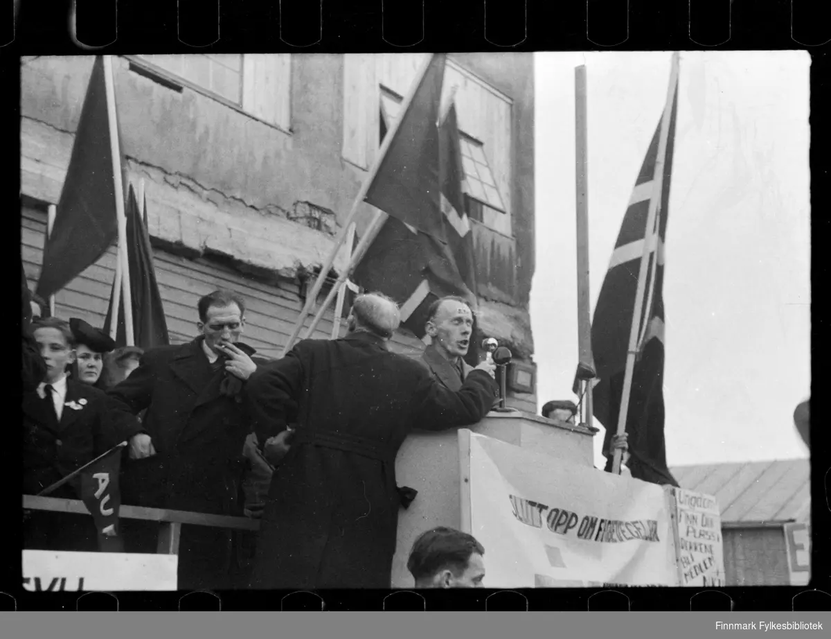 Foto av oppmøte ved betongskolen i Kirkenes, antagelig 1. mai 

Foto antagelig tatt på slutten av 1940-tallet, tidlig 1950-tallet 

Man kan se en mann i talerstolen. Ved siden av står en gutt som holder på et AUF flagg