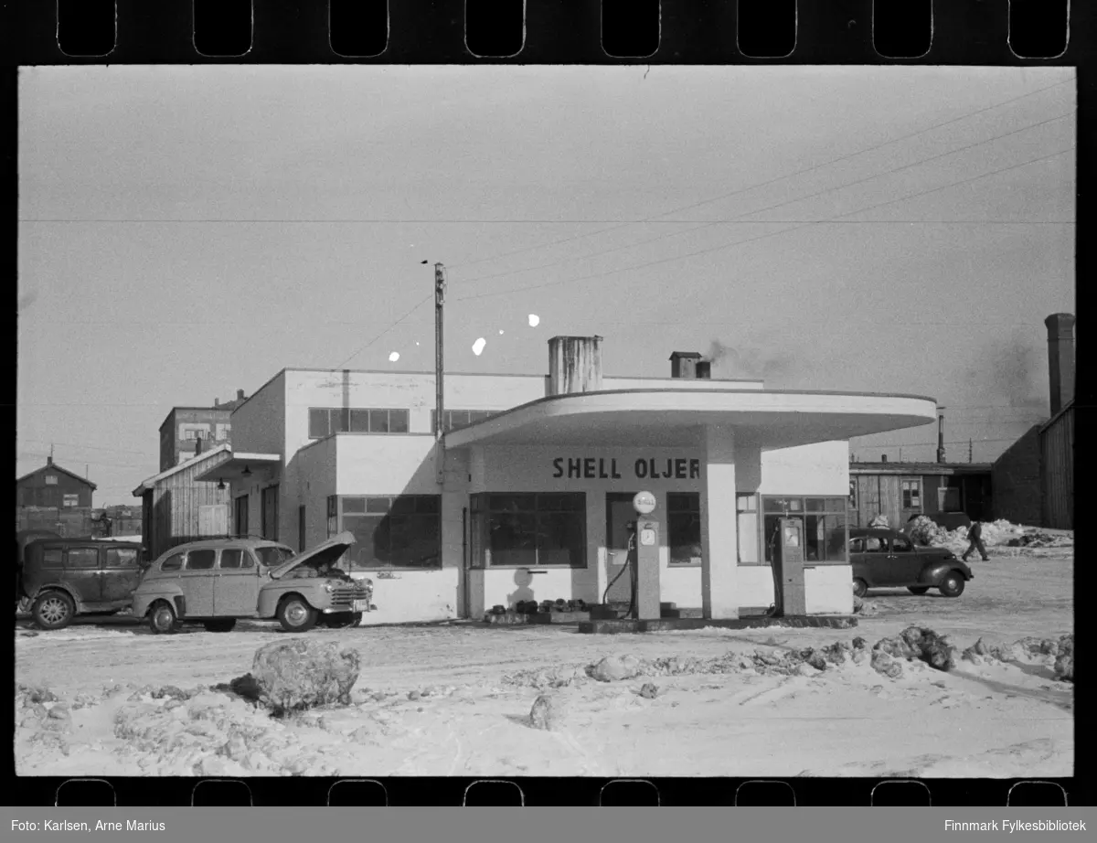 Foto av Shell Oljer bensinstasjon i Kirkenes 

I foto kan man se at gaten er en grusvei. Ved siden av bensinstasjonen ser man flere biler parkert og foran stasjonen er bensinpumper. Lys bil med panseret oppe er en Ford 1946-modell, reg.nr. Y-3300 (?) med undertekst DROSJE. Bak den en ca. 1930-modell.