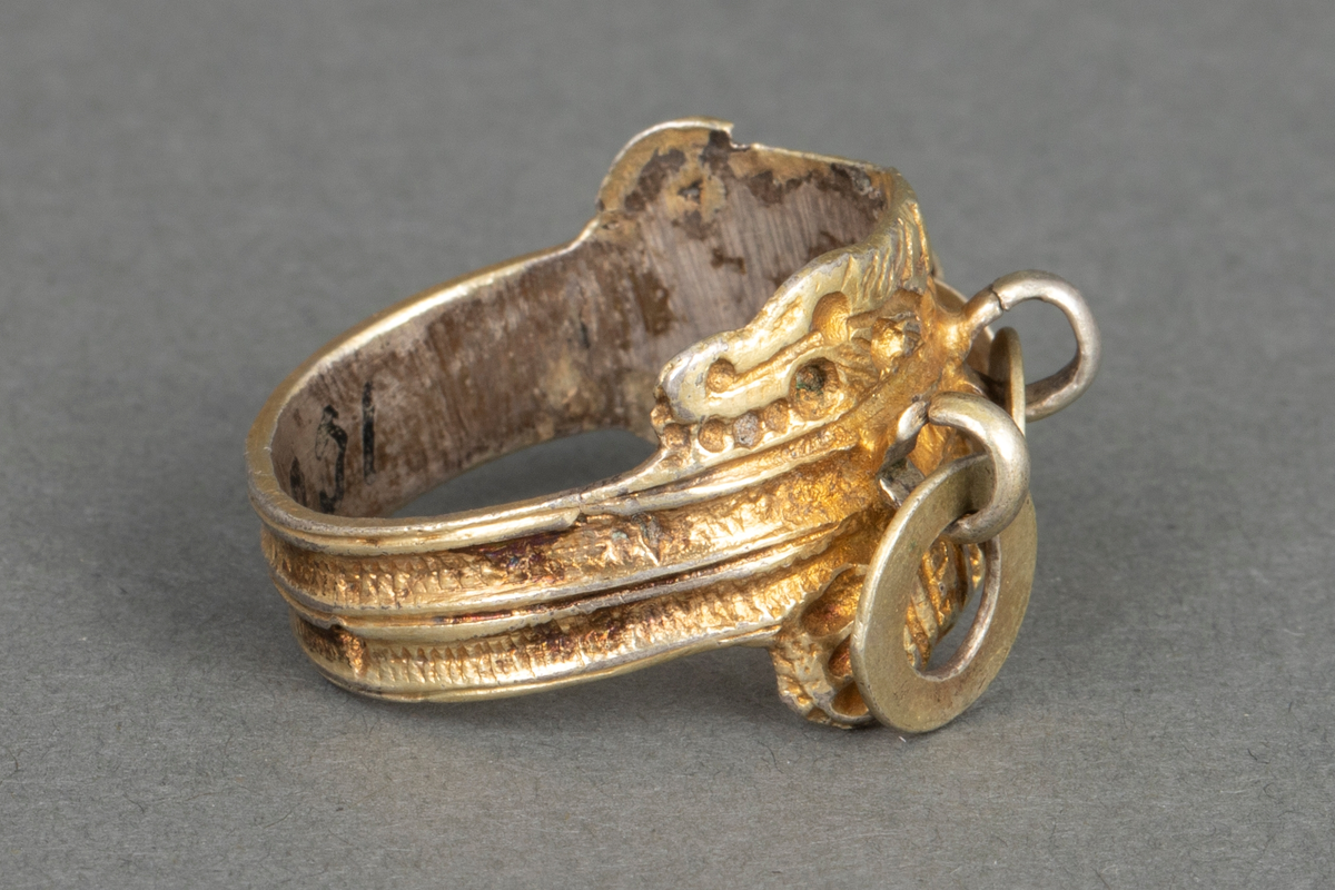 Støpt og siselert fingerring i forgylt sølv. Ringen er flat og bredest på forsiden. Den har "belter" (vulster) og stiliserte ormehoder, og på forsiden tre flate ringer festet i bøyler.