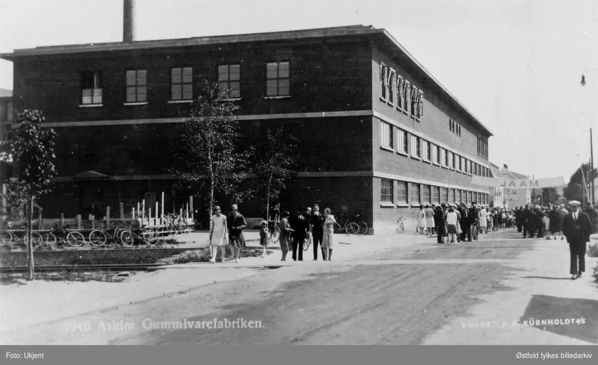 Askim gummivarefabrikk.  I forgrunnen sporet til den nedlagte Solbergfossbanen, til høyre i bakgrunn foregår et eller annet (innkomst for sykkelrace el. l.). 1920-åra. Postkort.