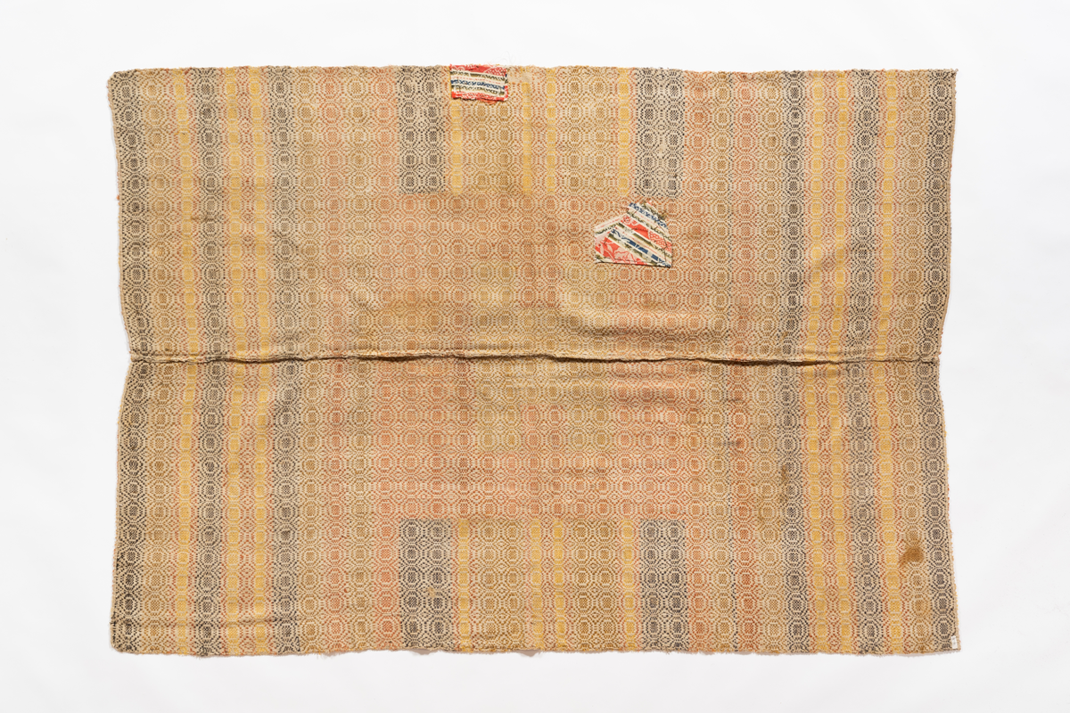 Täcke med varp av oblekt lin med inslag i brunt (gråblått enligt  katalogkorten), beige, gulrött och gult ullgarn. Två våder ihopsydda.