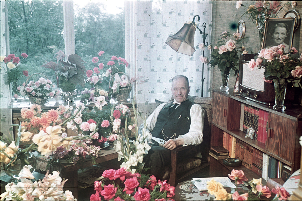 Fotograf Alf Schrøder fotografert på sin 70-års dag, omgitt av blomster. Alf Schrøder er fotografert sammen med en liten fugl som sitter på skulderen hans.