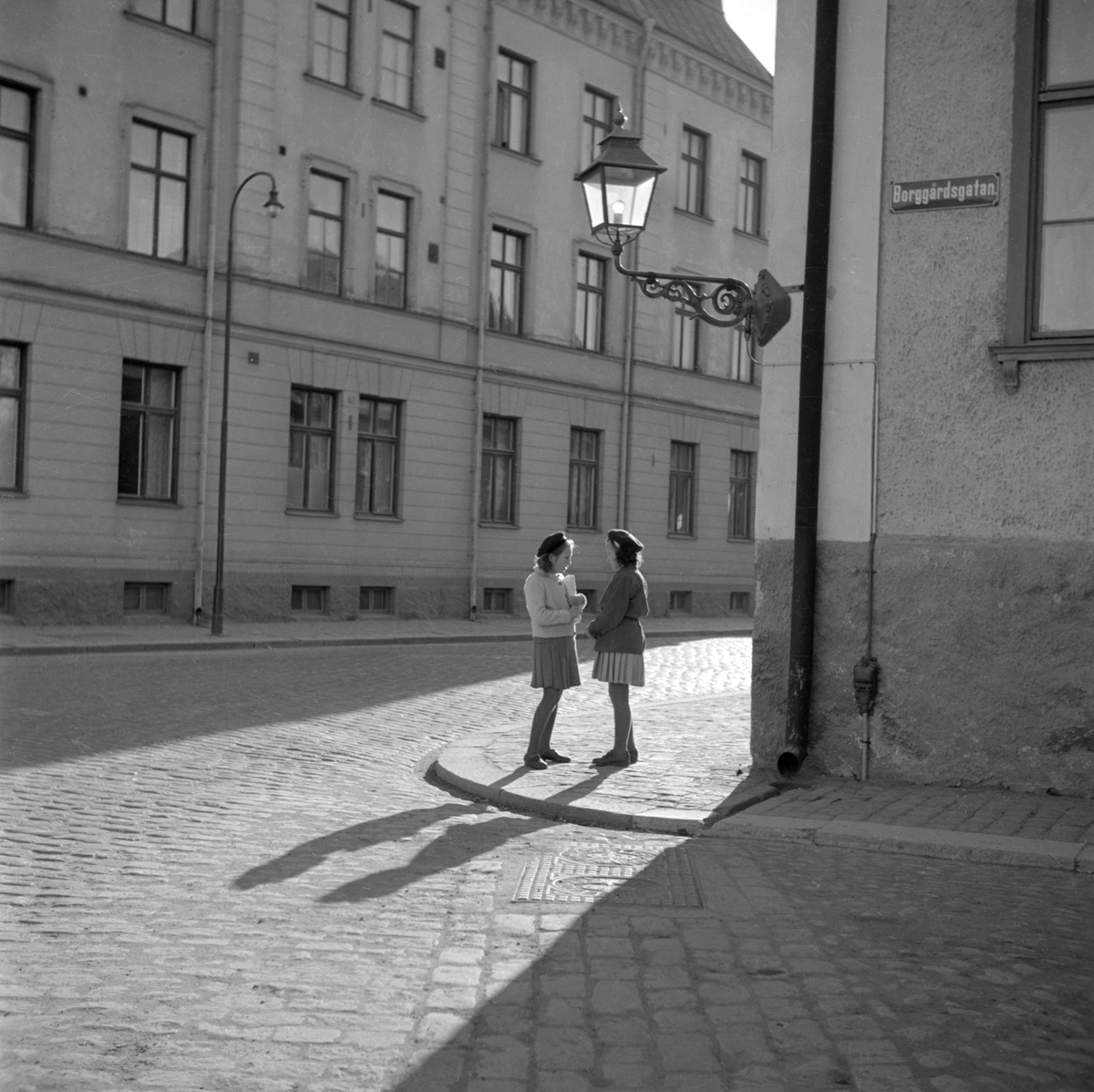 Unga väninnor råkas i hörnet av Storgatan-Borggårdsgatan i Linköping. Från väster slår den lågt stående solen skuggspel över platsen. Det pampiga Lorichska huset bildar fond och stiftets gamla domprostgård infattar motivet från motsatt håll. Bild från omkring 1950.