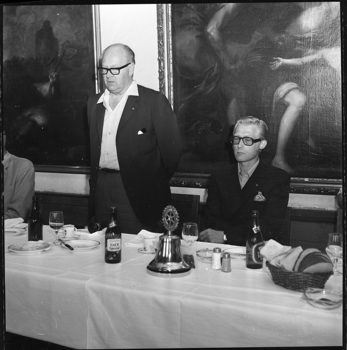 En man står upp vid ett matbord och håller sannolikt ett tal. På bordet en "Rotary-klocka", kaffekoppar, glas och ölflaskor. Han flankeras av sittande män.