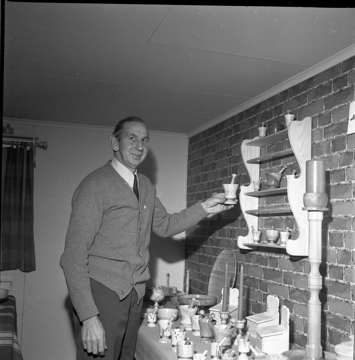 En man i cardigan vid ett bord och vägghylla med svarvade skålar och fat.