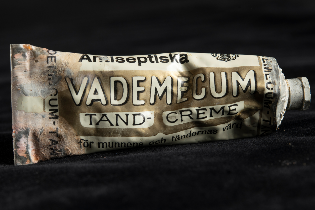 Metallförpackning innehållande tandkräm av märket Vademecum. Text i vitt och svart mot vit eller guldfärgad botten.