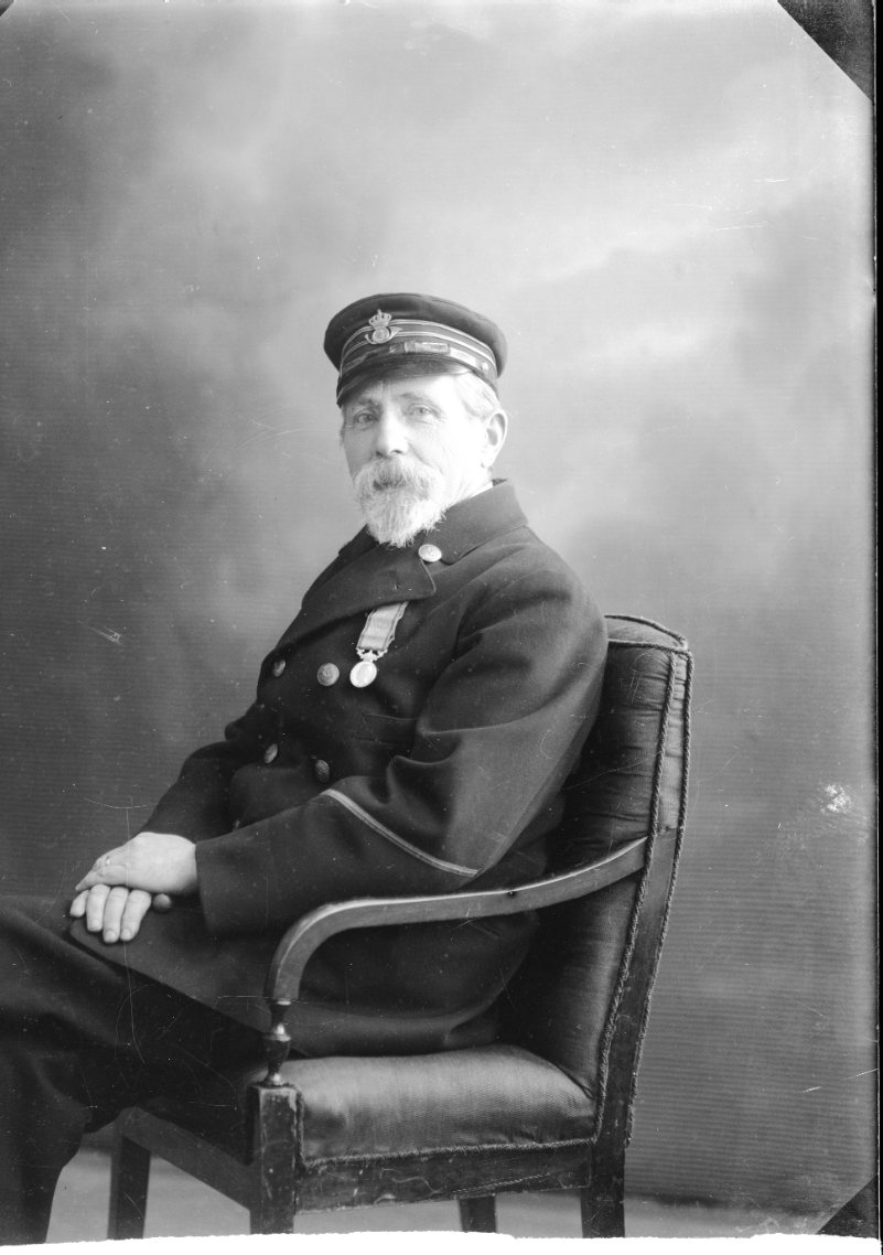 Bröstbild av en man med skägg som sitter på stol. Han bär postiljons uniform och keps, på bröstet hänger det en medalj - sannolikt "För nit och redlighet i rikets tjänst".