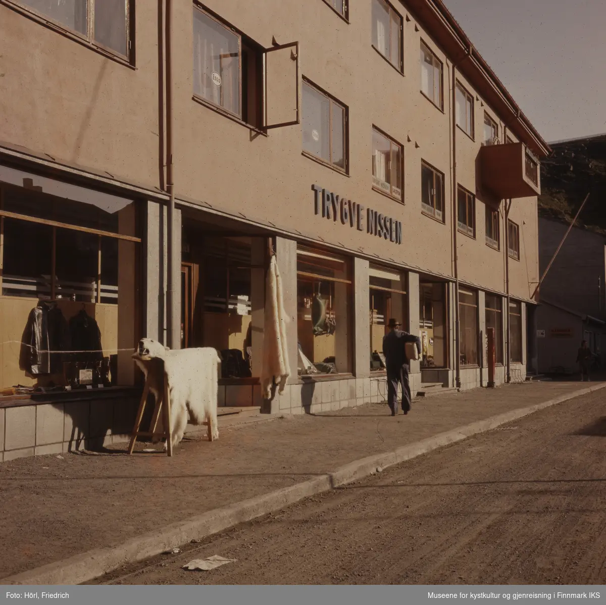 Butikken "Trygve Nissen" i Sjøgata i Hammerfest. I første etasjen er et vindu åpent. En Mann går i gata og bærer en pakke under armen. Foran butikken presenteres det en isbjørnskinn med hode.