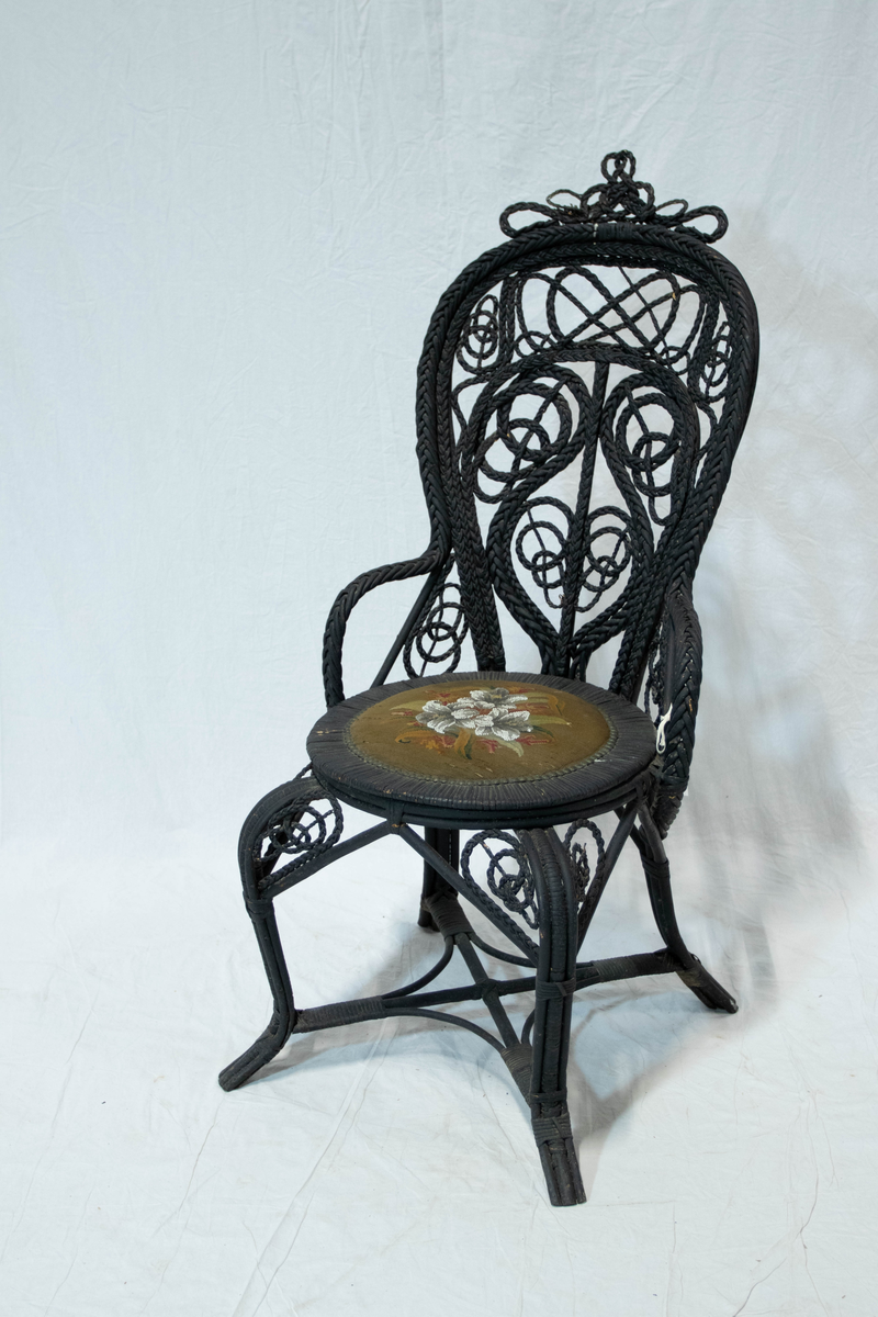 Svart kurvstol. Stolen har bøyde vidjeknipper som konstruktsjons ledd og et kunstferdig utført fletteverk av ornamental virkning i stolryggen. Setet er overstoppet med broderte perler og petit-point.