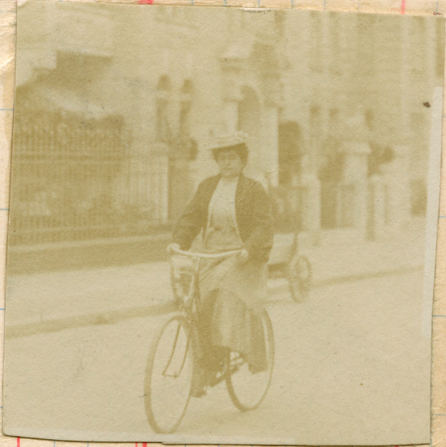 Motivet viser en kvinne med hatt og yttertøy som sykler på en vei. I bakgrunnen står en staselig bygård med smijernsgjerde.