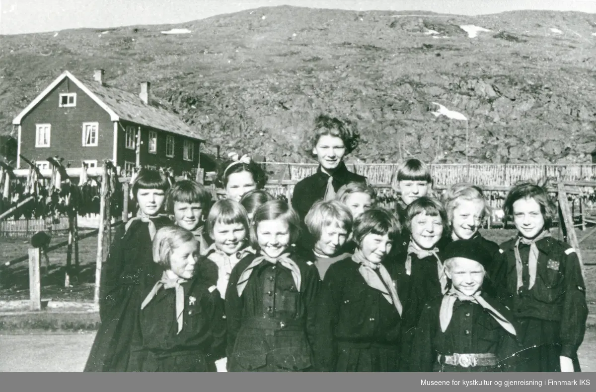 "Blåmeis". Gruppebilde av små jenter i speiderdrakt. I bakgrunnen ser man et hus og fiskehjeller. Antatt omkring 1939.