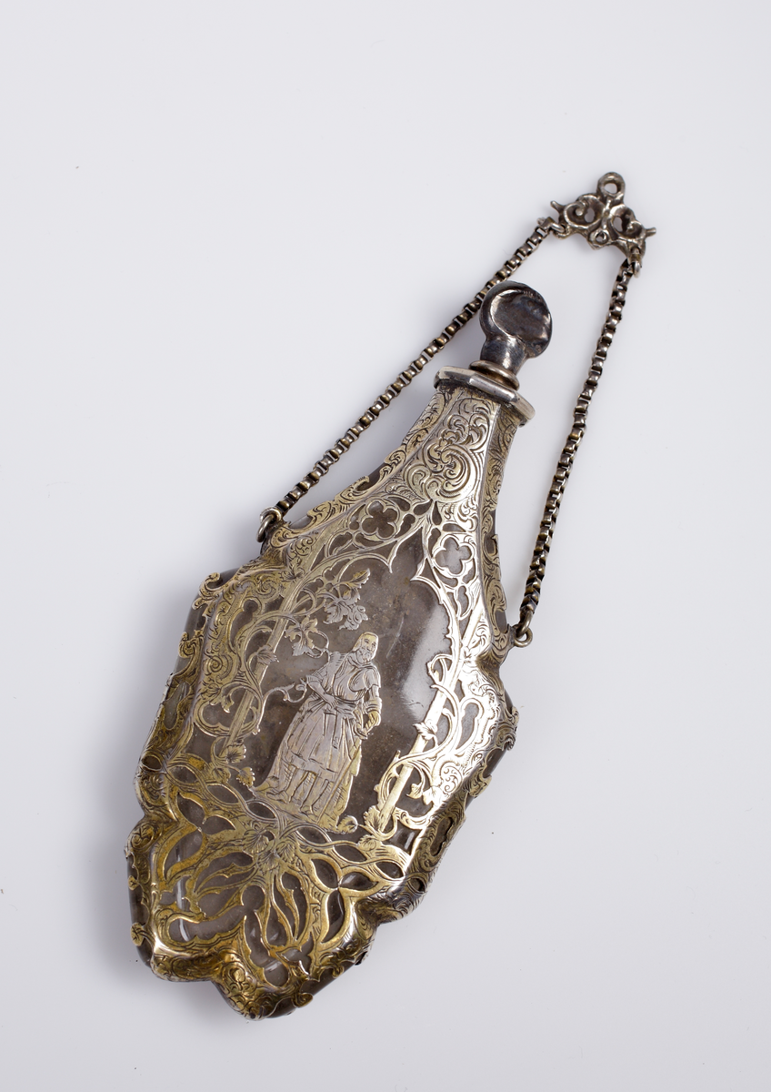 Platt parfymflaska (?) av kristall i skyddande förgylld och graverad silverinfattning, med liten kedja.

Trol. från Ludvig XV:s tid.

Inskrivet i huvudbok 1907.