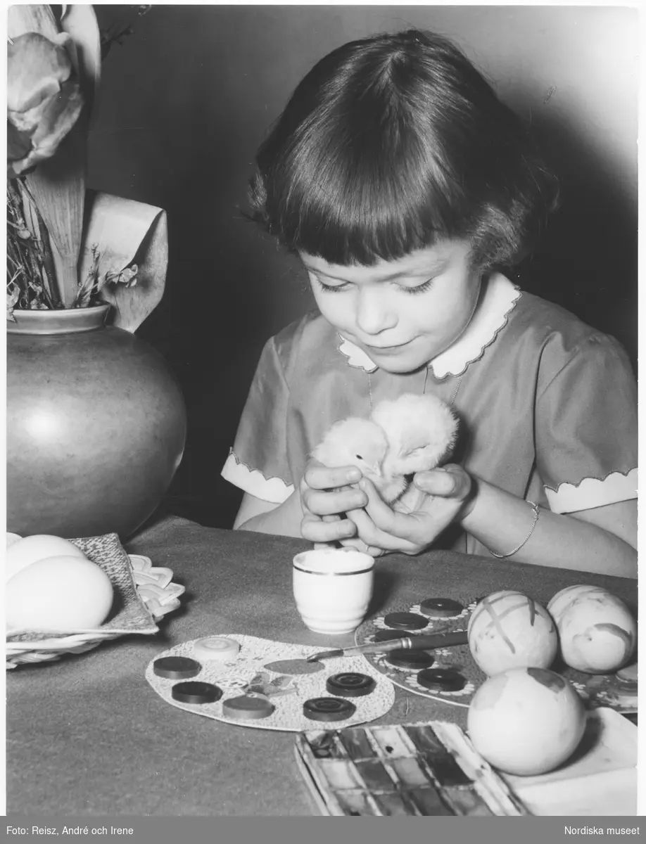 En flicka sitter vid ett bord med färg och målade ägg och håller två små kycklingar i sina händer.