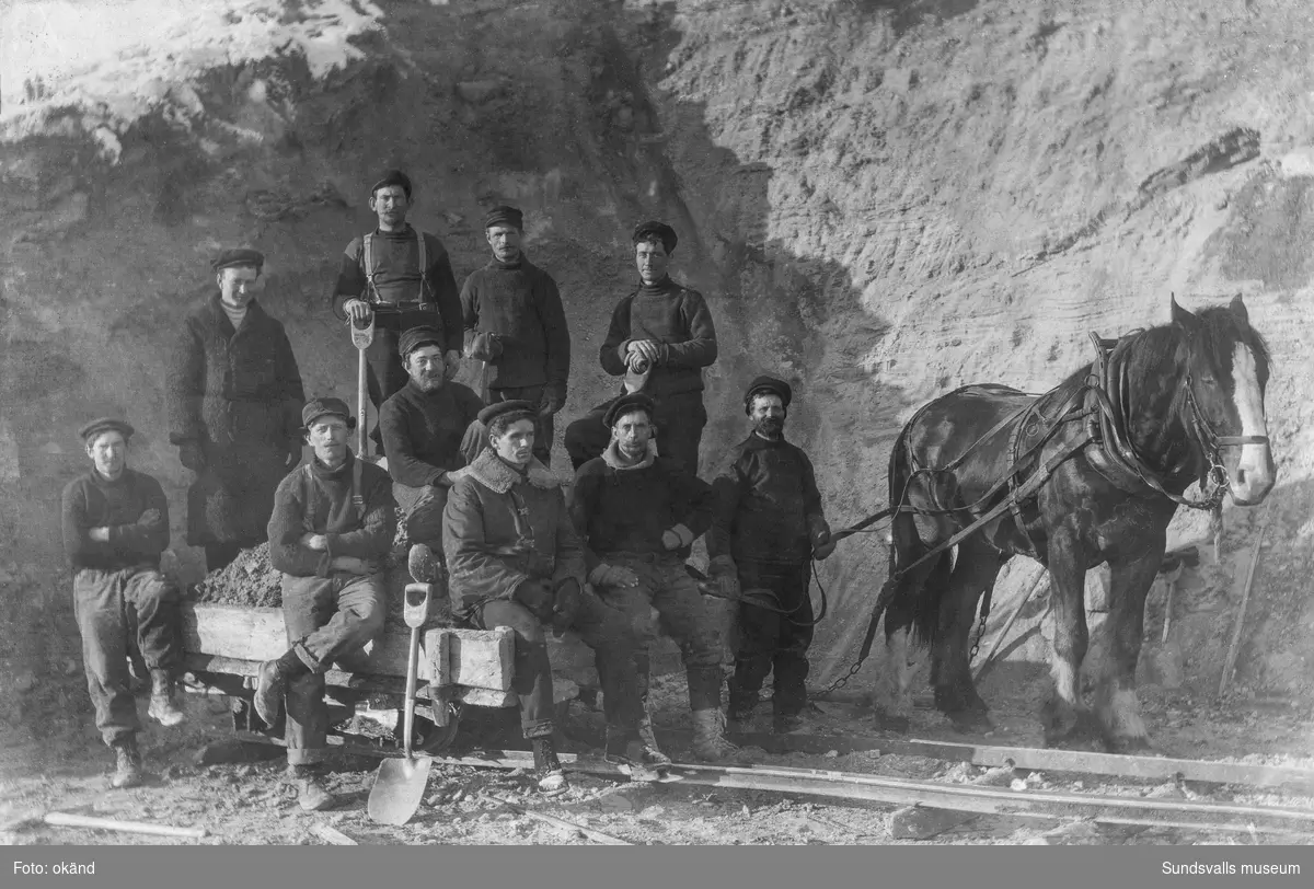 Gruppfoto med ett arbetslag på tio man som skottat en vagn på räls full med sand eller lera. Draghästen passar på att ta igen sig. Möjligen vid ett tegelbruk. Ur Anders Nylanders samling.