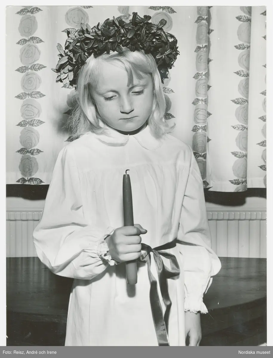 Lucia. En flicka klädd i Luciaklänning, med band i midjan och med en krans på huvudet håller ett ljus i handen.