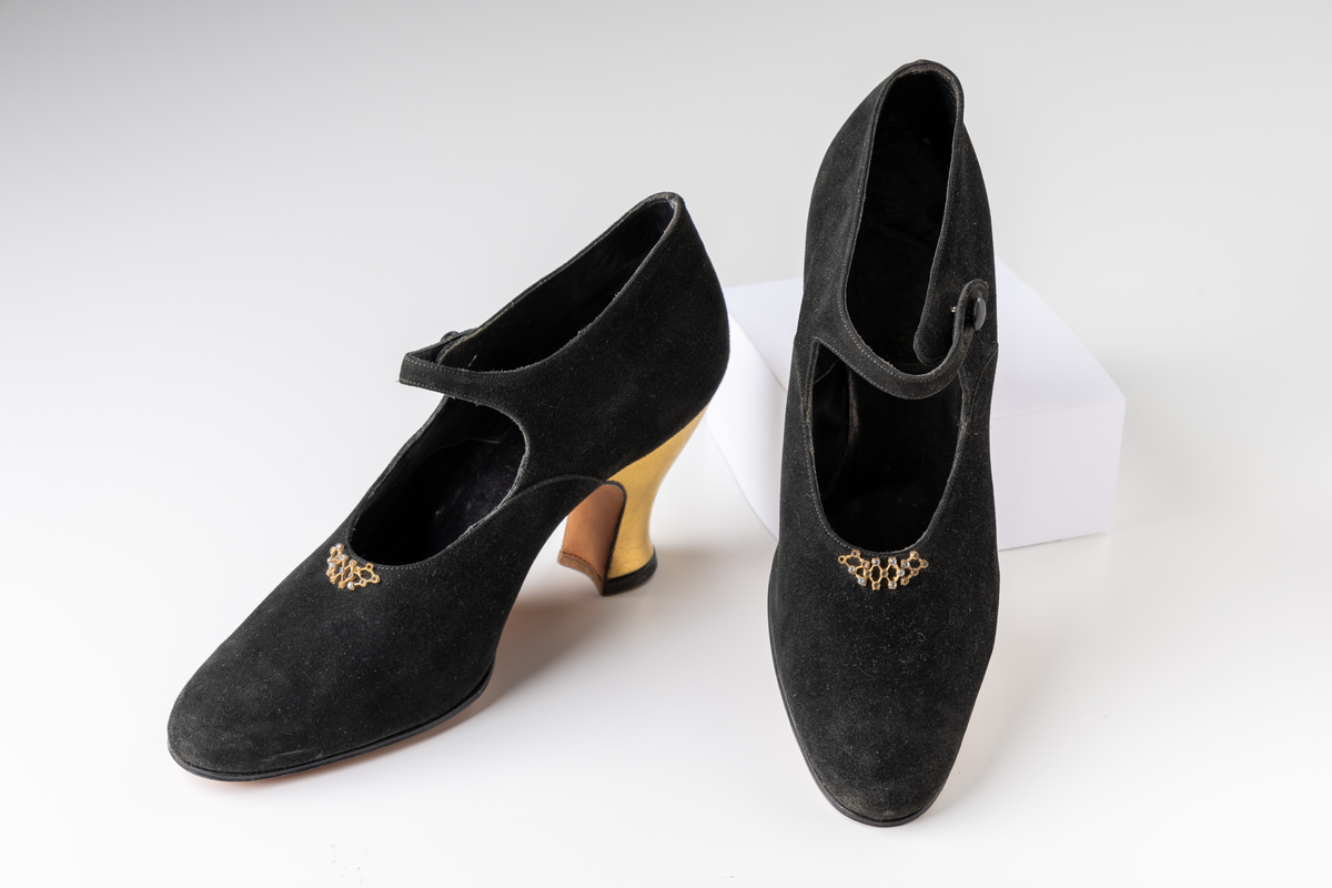 Ett par damskor från mitten av 1920-talet av svart mocka, klacken klädd med guldchevreau. Vristrem, som knäpps i sidan. Gulmetallspänne med vit strass monterad mitt på skon. Helfodrad. Lädersula. Storlek 36.