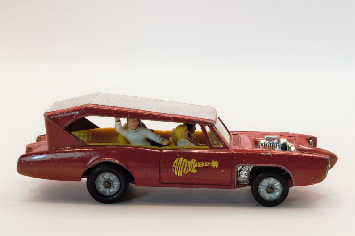 Rød lekebil fra 60/70-tallet. Produsert i Wales. The Monkeemobil. Modell av Dean Jeffries 1966 Pontiac GTO som var med i den amerikanske TV-serien The Monkees. Fire figurer i setene som spiller gitar og vinker, karakterene i serien spiller i band.