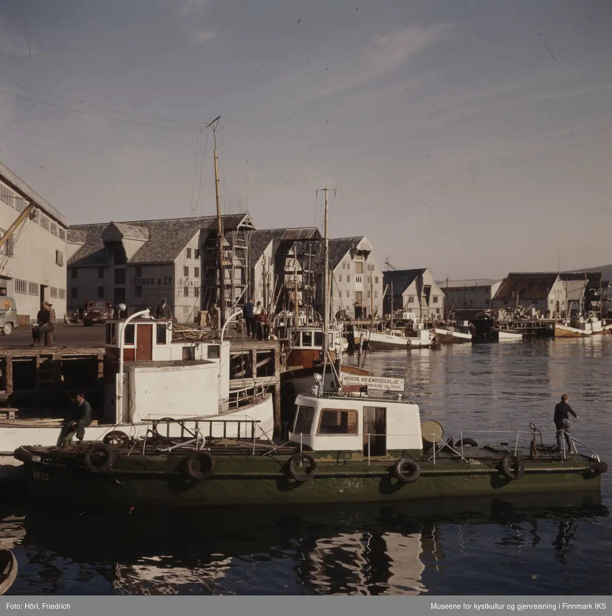 Havnesceneri i Tromsø i 1957 eller 1958. I forgrunnen ser man en grønn båt med et skilt for "Norsk Brændselsolje A/S". Ellers ser man fiskebåter ved kaia og mange pakkhus, blant annet til "Rieber & Co. A/S". Det ser ut som om det er flott vær og menn sitter på fiskebåten fremst i bilde samt på kaia.