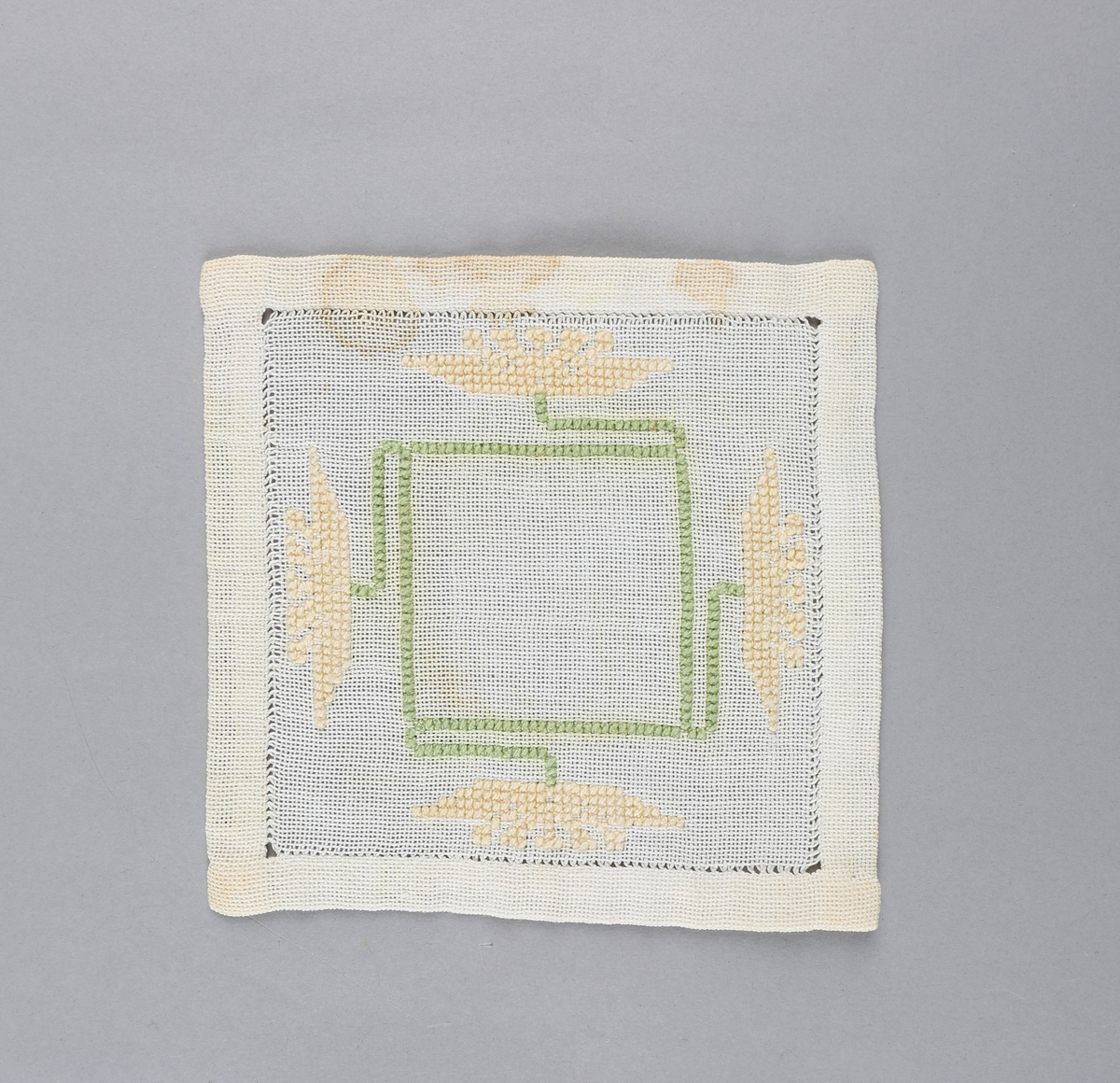 Tilnærmet kvadratisk dekkebrikke i hvitt tekstil med brodert, grønt kvadrat med fire stilker i sidene med gule blomster i endene.
