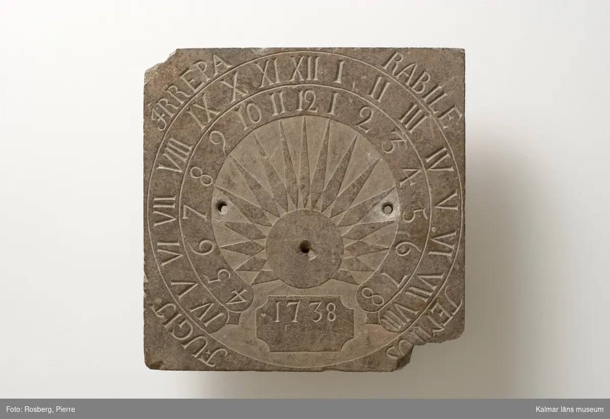 KLM 7329. Solur. Tillverkat av rödaktig sten. Fyrkantig. Både romerska och arabiska siffror i cirkel. Inskrift: årtalet 1738 samt Fugit irreparabile tempus.