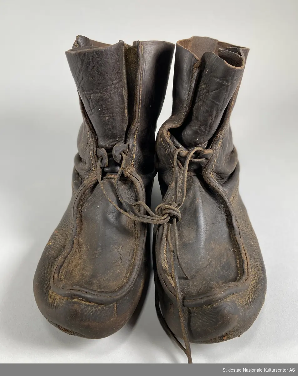 Et par skinnsko kalt "lauparsko?" med skinnreimer (snøring) for å feste rundt skoene. Snøringen er sydd fast mellom to sømmer i skaftet. Sålelær er sydd sammen med overlæret. Skoene ser ut til å ha hatt såler, men disse er fjernet (tydelige spor etter fjerning). Uforet sko. Størrelse 45.