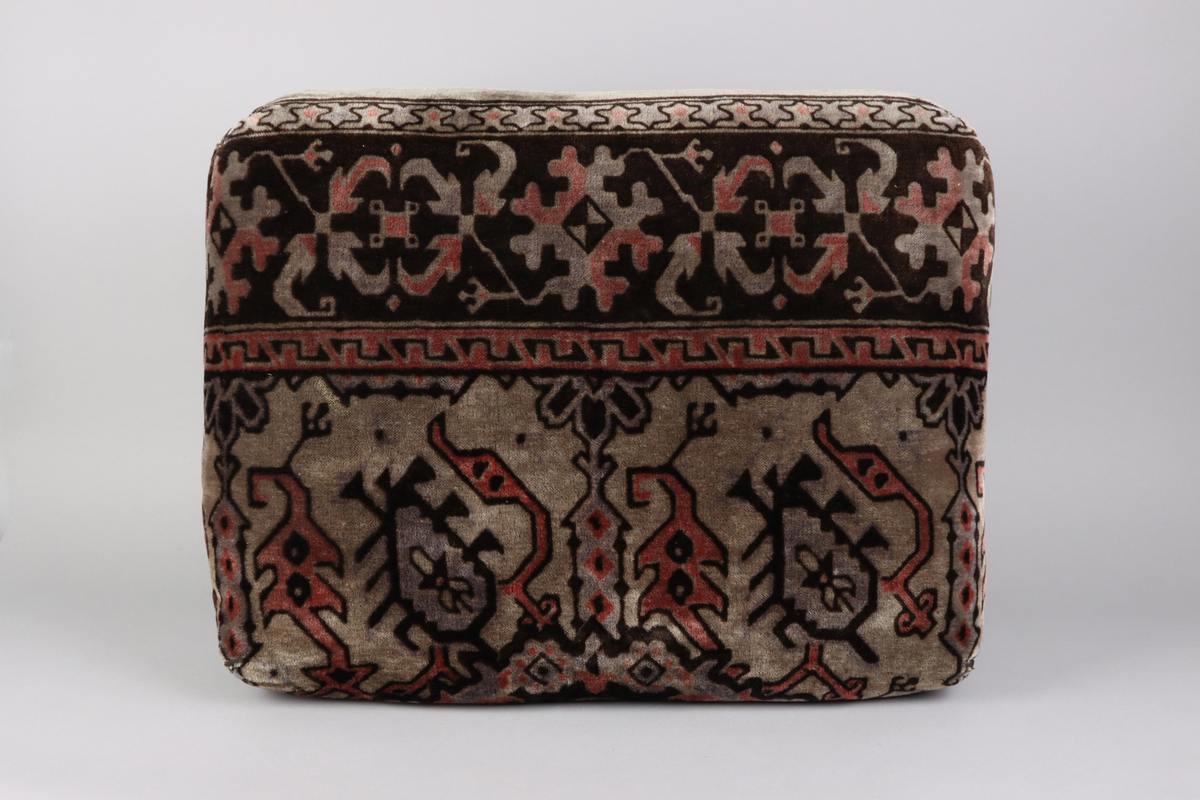 En kudde av mönstrad schagg. Orientaliserande mönster i färgerna grå, svart och vinröd. Del i en grupp med överkast och kuddar till en sk. Ottoman, en typ av bäddsoffa.
