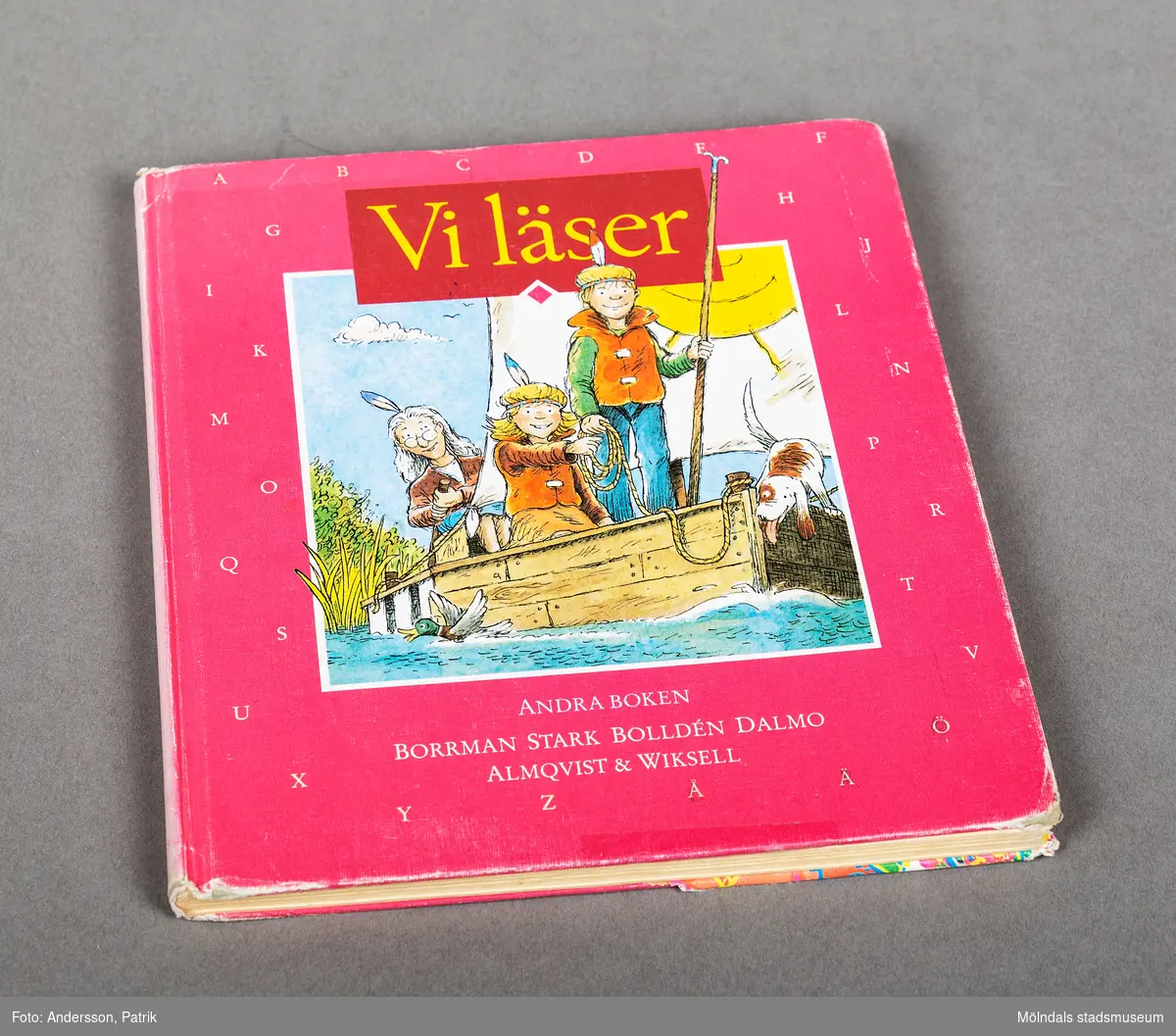 Bok: Vi läser. Andra boken. På framsidan finns en teckning av en äldre kvinna, två barn och en hund. De seglar med en flotte. Omslaget har rosa bakgrund med vita bokstäver. 

Boken innehåller enklare texter av flera olika författare och illustratörer.