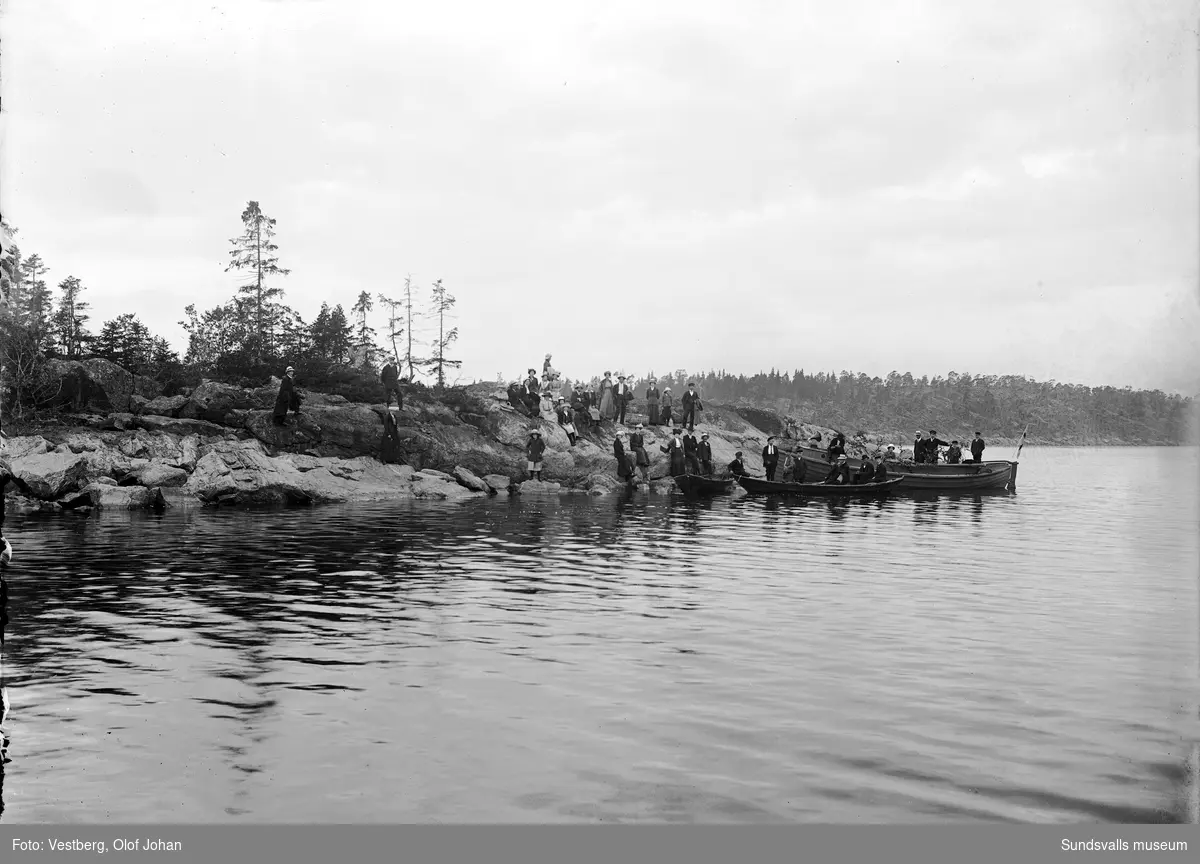 Båtutflykt till ön Skigan i Juniskärs skärgård. Tre båtar har lagt till vid en udde och ett större sällskap står på klipporna med bland annat fikakorgar. Den större båten är lövad och i fören står en trattgrammofon.