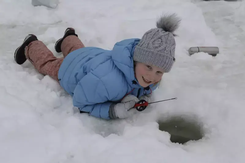 En jente med vinterklær ligger på isen og pilker med en rød pilkestikke