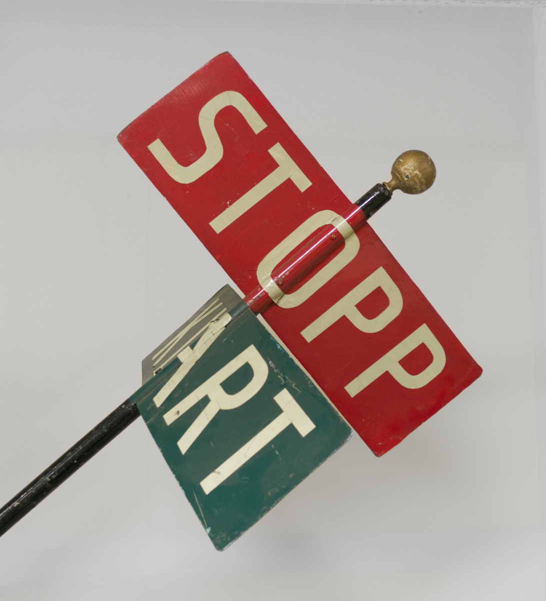 To rektangulære metallskilt. Et rødt skilt med teksten STOPP og et grønt skilt med teksten KLART. Skiltene er montert i hver sin retning på en høy stang med håndtak, slik at skiltene enkelt kan dreies rundt.