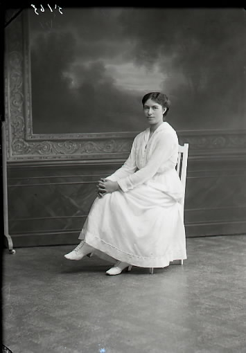 Ateljébild. Kvinna sittande i helfigur, troligen helt klädd i vitt, även snörkängorna. Beställare: Fröken Kristensson, Hotellet, Falkenberg.