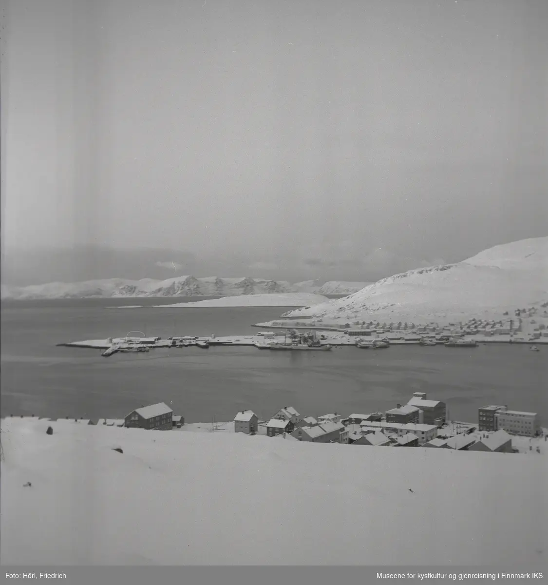 Bildet viser utsikten over Hammerfest og bydelen Fuglenes om vinteren. I forgrunnen ser man ulike bygg i sentrum og på Hauen, blant annet hotellet. I bildemidten ser man bydelen Fuglenes med slippen og et skip som ligger til kais. Takene er snødekket på gjenreisningshusene på Rosmolla og bak der, på Meland, står det fiskehjeller. I Sørøysundet ligger Melkøya og Sørøya lengst bak i bilde.