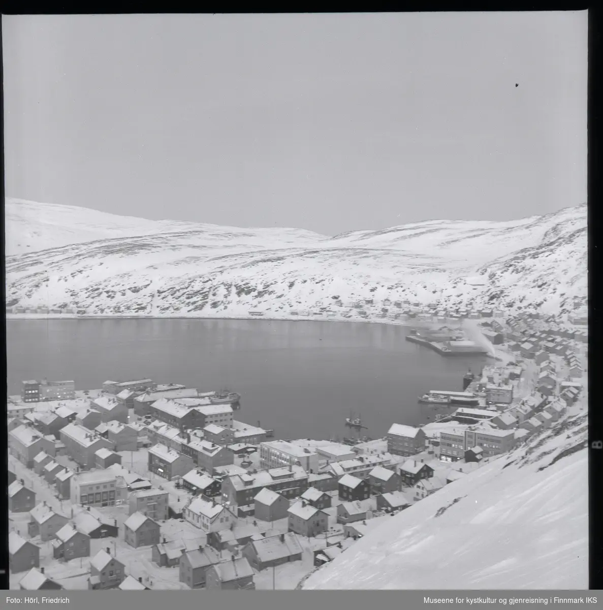 Utsikt over Hammerfest sentrum fra Salenfjellet i vinteren 1957/1958. Mesteparten av byen er gjenreist og man ser mange sentrale bygg som kommunehuset, Handelshuset Nissen, rådhuset, bankbygget, Findusfabrikken og Hauans materialhandel. I bakgrunnen ligger bydelen Molla og det snødekkede fjellandskapet.