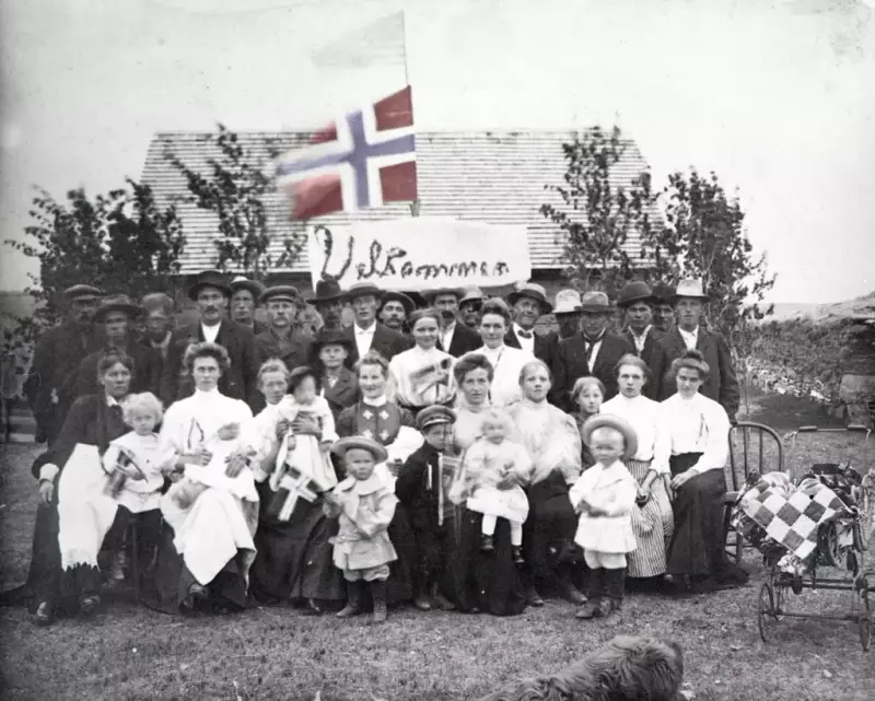 A group of women, men and children sit outside a small house. Small trees on either side of the house and an empty pram stand to the right of the group. Above the group is a sign with the inscription Welcome and a flagpole with first a Norwegian flag, and then an American flag at the top. Some children also hold small Norwegian flags in their hands.