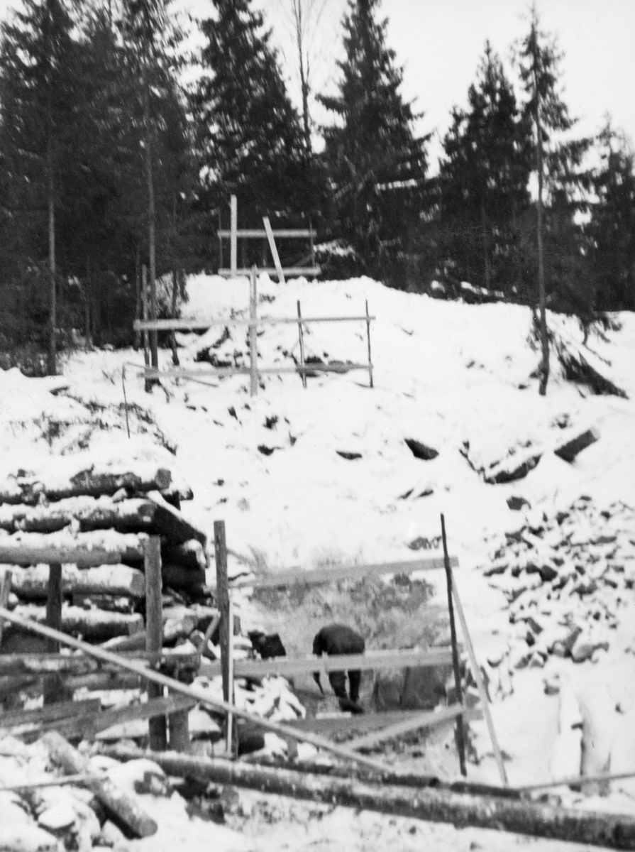 Fra rivinga av den gamle dammen ved Sølvstufossen, øverst i Ågårdselva i Tune i Østfold, som er en del av Nedre Glommas vestre løp. Dette arbeidet ble gjort vinteren 1936, i en periode da det knapt rant vann i elveløpet, samtidig som man startet med foreskaling til og støping av en ny dam like ovenfor. Vi ser rester av den forrige dammen, som var en steinfylt tømmerkistekonstruksjon, til venstre i forgrunnen. Rett framfor fotografen sto den arbeider bøyd over noe han arbeidet med i sjølve elveløpet. Ågårdselva er et cirka fem og en halv kilometer langt vassdrag som renner fra den nordvestre enden av Isnesfjorden (Vestvannet) i Nedre Glommas vestre løp, sørvestover gjennem berglendt terreng mot innsjøen Visterflo. Høydeforskjellen mellom Isnesfjorden og Visterflo er på bortimot 25 meter. Ågårdselva har tre fossefall, det øverste her ved Sølvstu, deretter ved Valbrekke og nederst ved Solli. Behovet for den dambygginga vi ser på dette fotografiet var forårsaket av selskapene Borregaards og Hafslunds inngrep i Glommas østre løp, hovedløpet, som i lavvannsperioder gjorde det svært vanskelig å få tømmer som skulle til bedrifter nedenfor Sarpsfossen via Mingevannet, Isnesfjorden og tømmertunnelen til Eidet gjennom det trange sundet ved Trøsken. Dette forsøkte man å løse ved å heve vannspeilet i Isnesfjorden med en dam som skulle plasseres 10-15 meter ovenfor den eksisterende dammen ved Sølvstufossen. Se mer informasjon under fanen «Opplysninger».