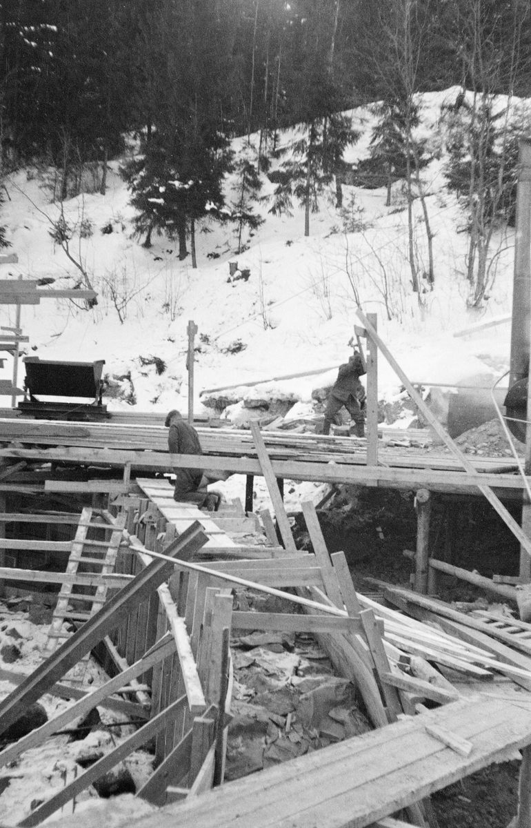 Fra bygginga av en ny dam ved Sølvstufossen, øverst i Ågårdselva i Tune i Østfold, som er en del av Nedre Glommas vestre løp. Anleggsarbeidet startet vinteren 1936, i en periode da det knapt rant vann i elveløpet. Dette fotografiet viser ei av forskalingene som ble bygd med sikte på å kunne støpe en mur på tvers av elveløpet. Vede elvebredden i bakgrunnen ser vi ei skinnegående vogn på en trallebane, som antakelig ble bygd for å oppnå effektiv transport av redskaper og materialer til og fra byggeplassen. Ågårdselva er et cirka fem og en halv kilometer langt vassdrag som renner fra den nordvestre enden av Isnesfjorden (Vestvannet) i Nedre Glommas vestre løp, sørvestover gjennem berglendt terreng mot innsjøen Visterflo. Høydeforskjellen mellom Isnesfjorden og Visterflo er på bortimot 25 meter. Ågårdselva har tre fossefall, det øverste her ved Sølvstu, deretter ved Valbrekke og nederst ved Solli. Behovet for den dambygginga vi ser på dette fotografiet var forårsaket av selskapene Borregaards og Hafslunds inngrep i Glommas østre løp, hovedløpet, som i lavvannsperioder gjorde det svært vanskelig å få tømmer som skulle til bedrifter nedenfor Sarpsfossen via Mingevannet, Isnesfjorden og tømmertunnelen til Eidet gjennom det trange sundet ved Trøsken. Dette forsøkte man å løse ved å heve vannspeilet i Isnesfjorden med en dam som skulle plasseres 10-15 meter ovenfor den eksisterende dammen ved Sølvstufossen. Se mer informasjon under fanen «Opplysninger».