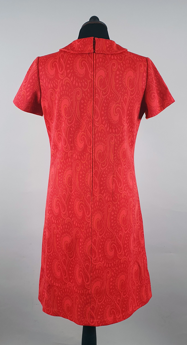 Rød kjole med orientalske mønster. Rett fasong med korte ermer og plisséfolder midt foran. Skjortekrage og glidelås i ryggen.