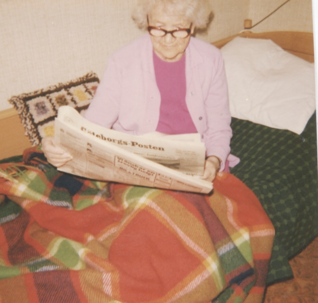 En okänd kvinna (boende?) sitter på en säng och läser Göteborgs-Posten, Brattåshemmet 1970 - 80-tal. Kvinnan har en filt över benen. I bakgrunden ses ett par kuddar.
